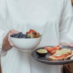 Person holding a bowl of fresh berries and a plate with avocado toast, displaying a healthy breakfast spread.